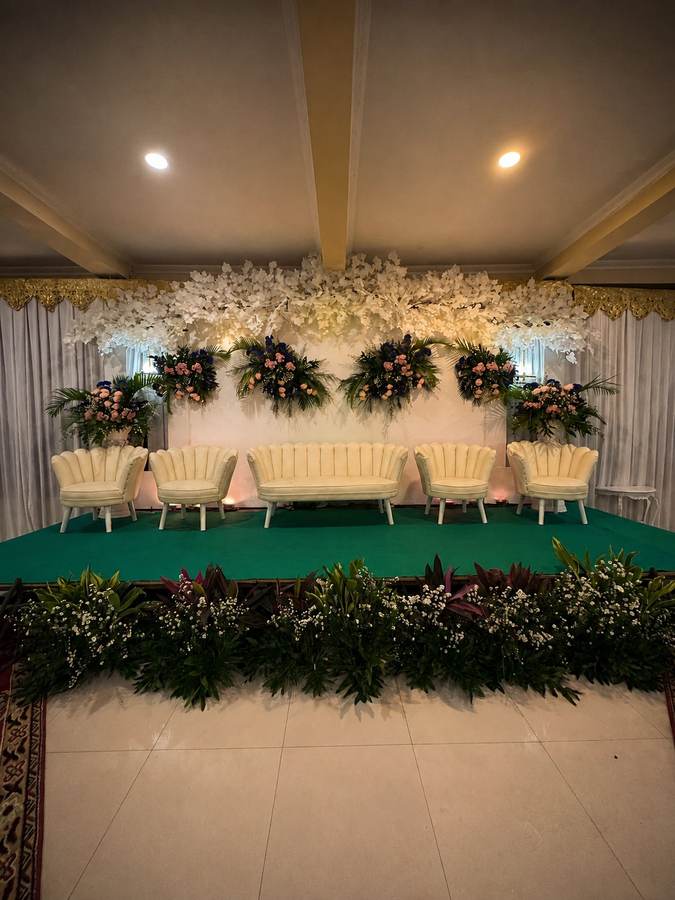 Pelaminan dengan sofa krem dan backdrop bunga di atas panggung