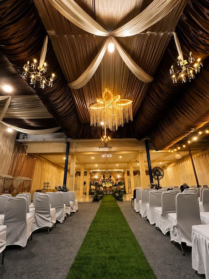 Interior Event Hall Eyang Akung: plafon drapery cokelat keemasan, lampu gantung kristal, dan lorong karpet hijau menuju pelaminan dengan kursi tamu tertata di kedua sisi